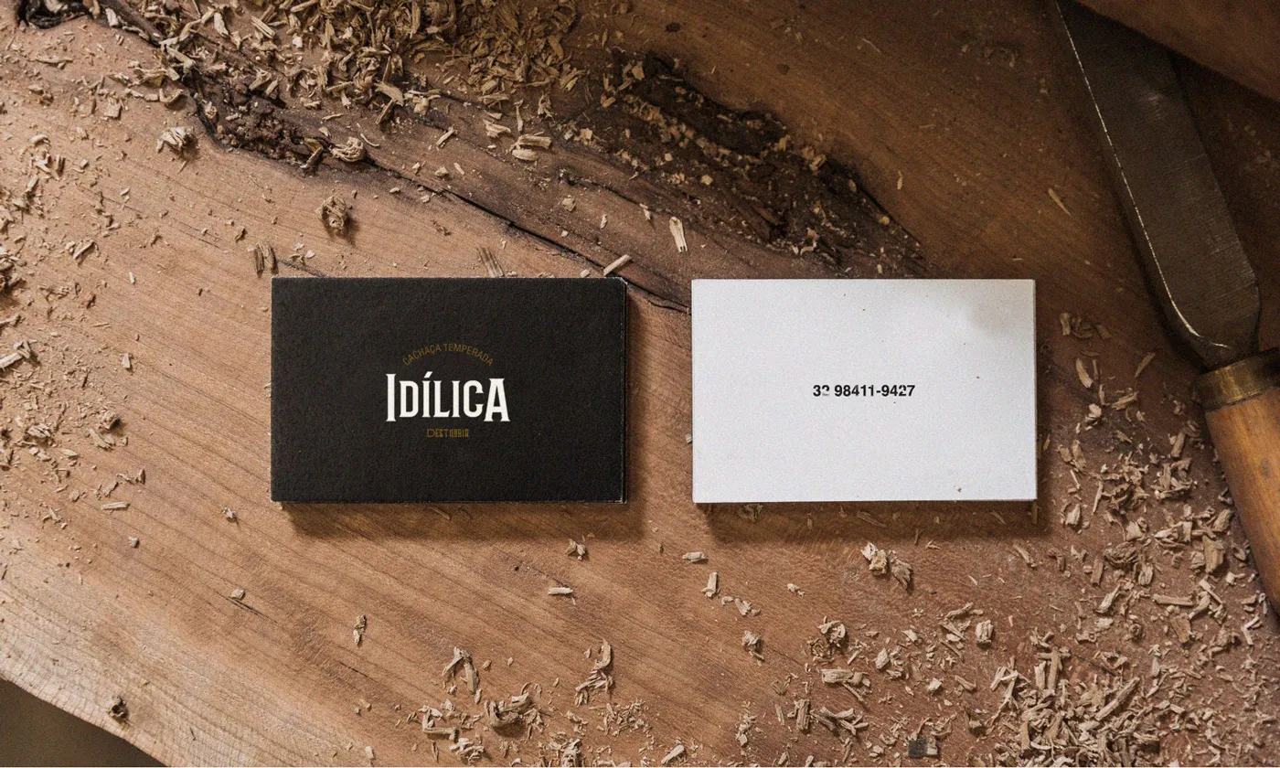 Cartão de visitas, um lado preto com o logo o outro branco com telefone. Dispostos sobre mesa de madeira cheia de pedaços de serragem