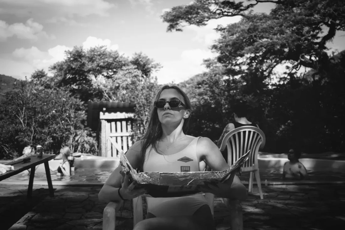 Black and white photo of a girl sun bathing. front angle shot. she is sitting in a chair with sunglasses and there is a pool on the background