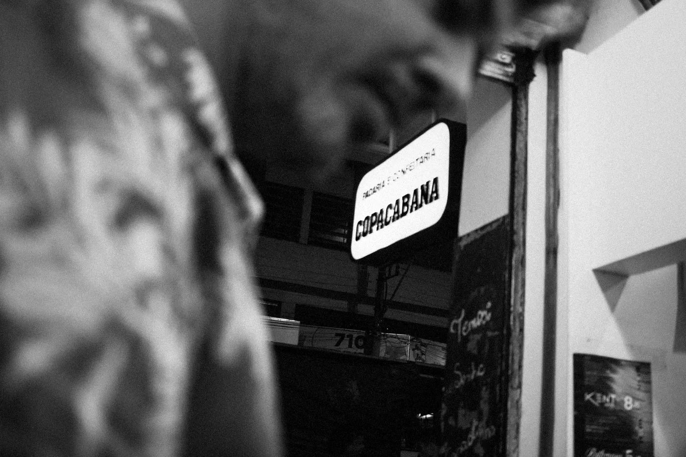 A blurred man on front. A Sign of a bakery with COPACABANA writen on it. Shot in black and white
