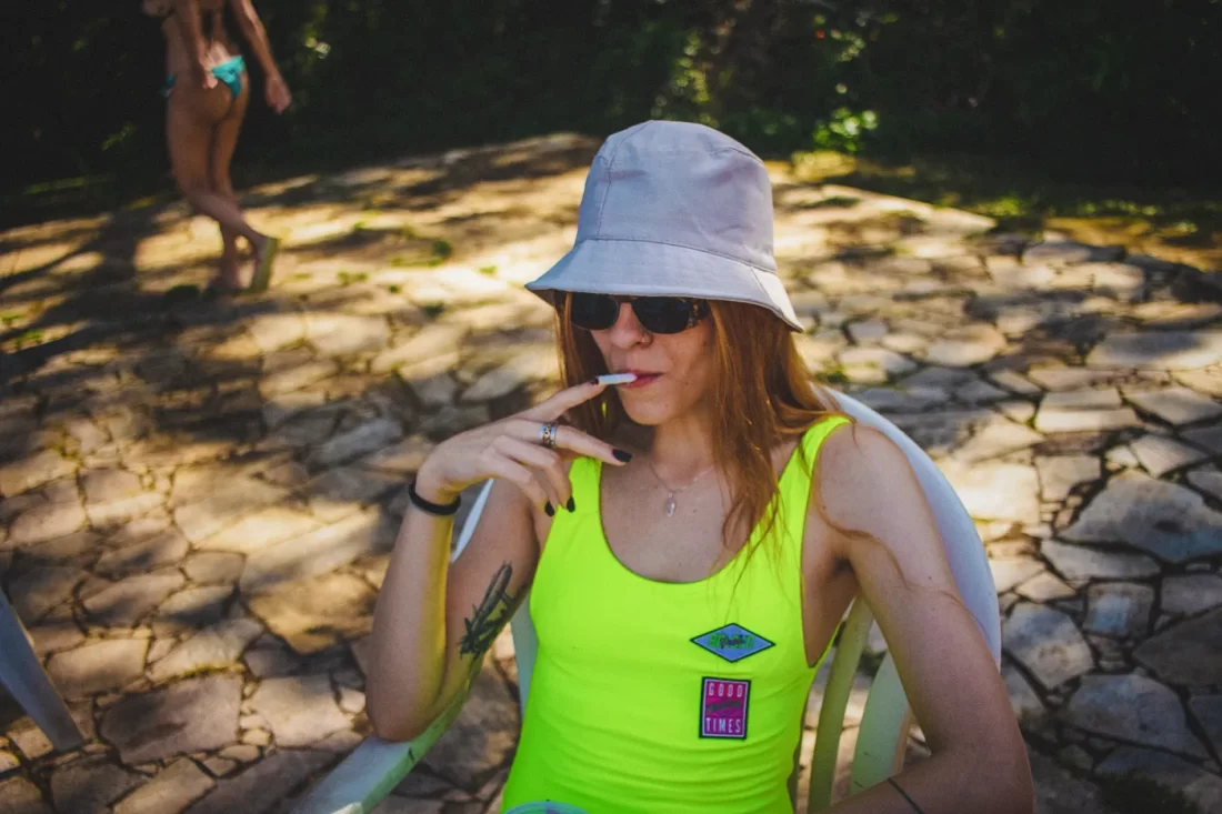 Redred woman smoking by a pool. She is wearing a lime bathingsuit, sunglasses and a white hat