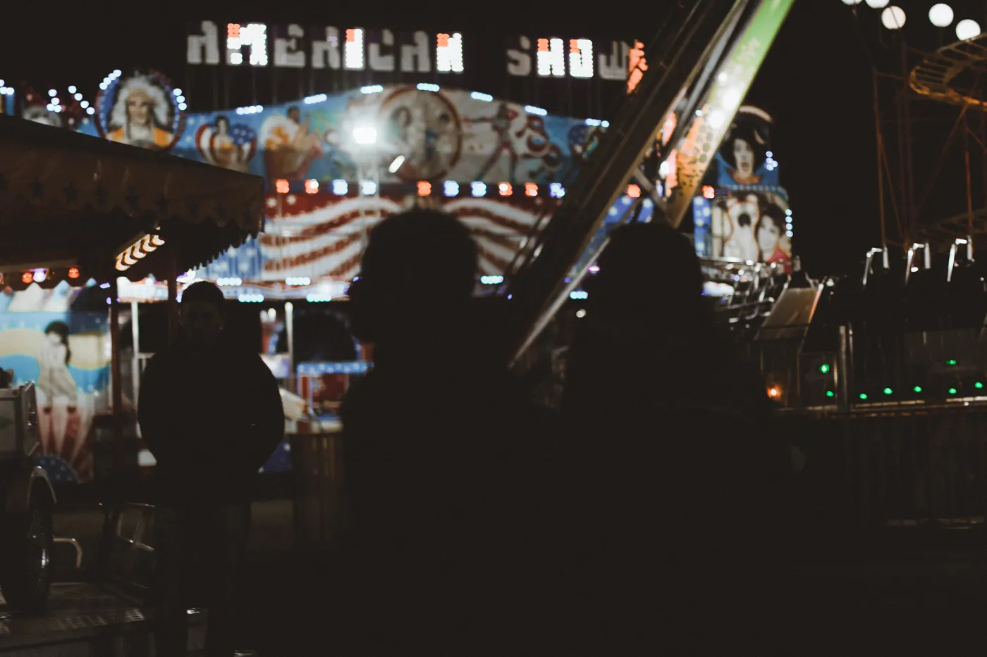 Nighttime Urban Photography – Silhouettes and Carnival Lights by Rodrigo Baumgratz
This nocturnal photograph by Rodrigo Baumgratz captures dark human silhouettes set against the harsh neon of a carnival ride titled “American Show.” The visual tension between shadowed figures and overexposed amusement park lights creates a powerful image of distance, spectacle, and identity. Ideal for portfolios centered on urban photography, documentary aesthetics, or visual commentary on mass entertainment, the photo blends mood and observation with a cinematic edge.  