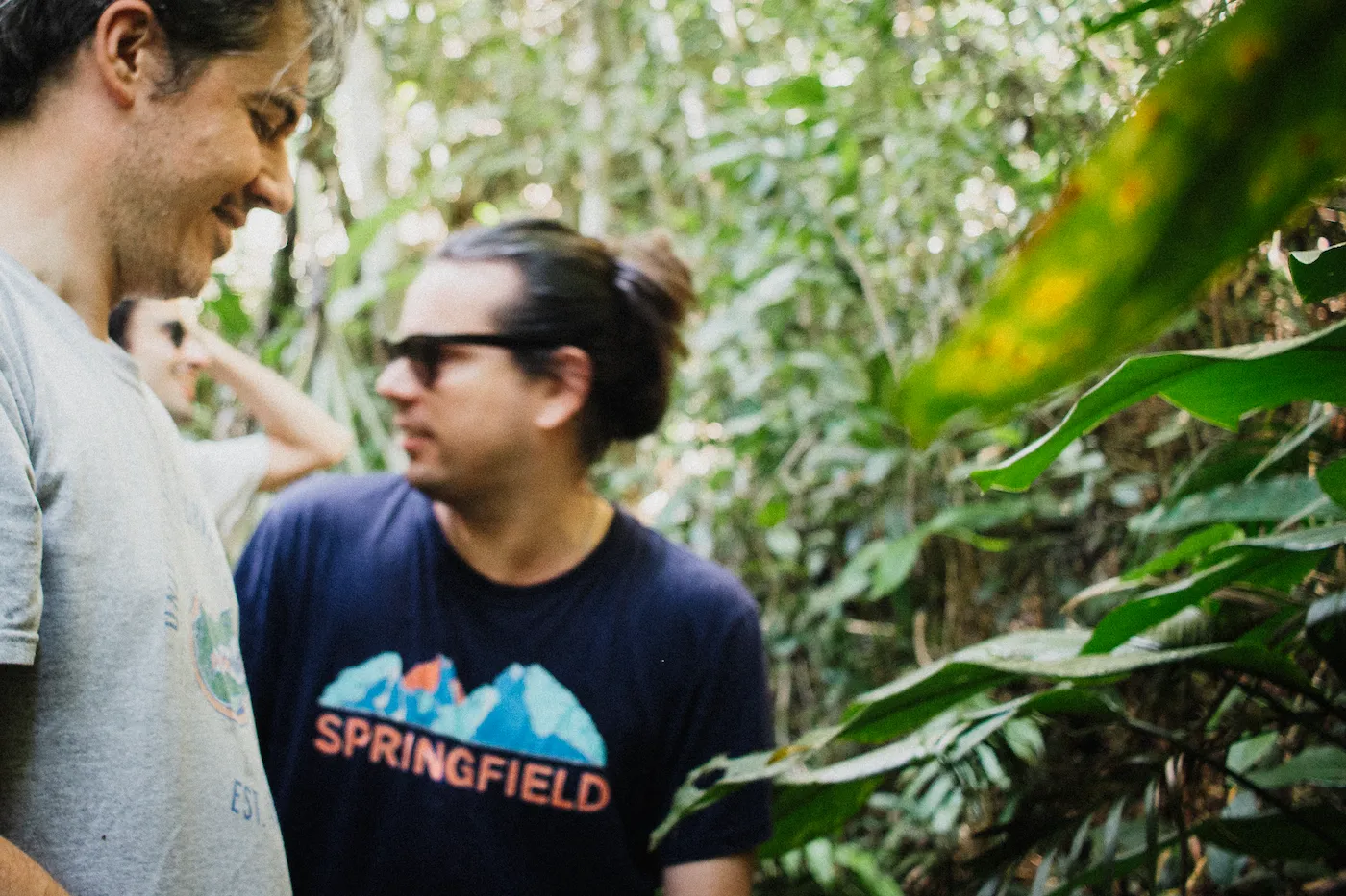 3 guys on the middle of the jungle. One, wearing gray on front, blurred. The second one, focused, wears sunglasses and a tee with springfield writen on it. The third one, on the back, holds his head and laught