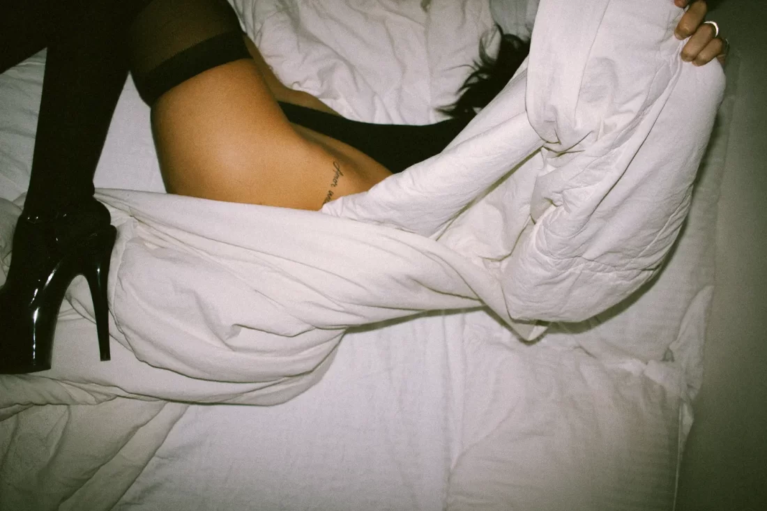 Uma mulher em uma cama tampando o rosto com o edredon branco. Ela usa um body com meia calça e salto preto