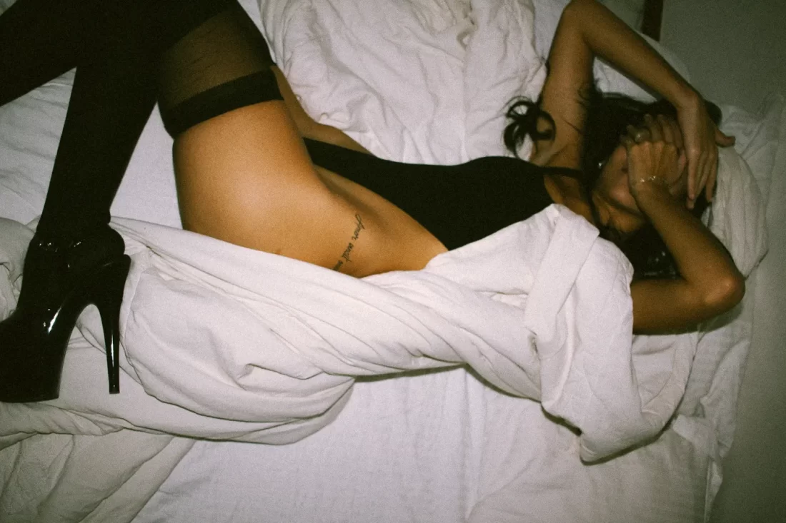 Uma mulher em uma cama tampando o rosto. Ela usa um body com meia calça e salto preto