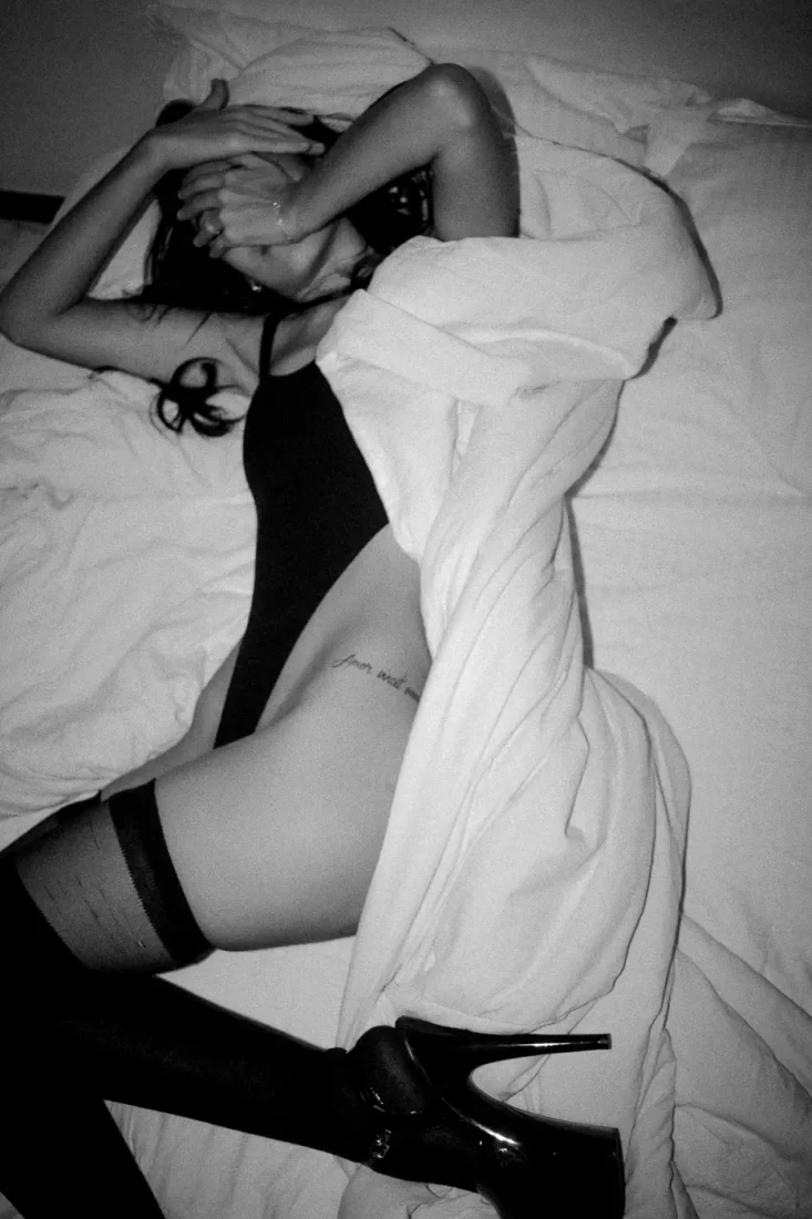 Uma mulher em uma cama tampando o rosto. Ela usa um body com meia calça e salto preto. Foto em preto e branco