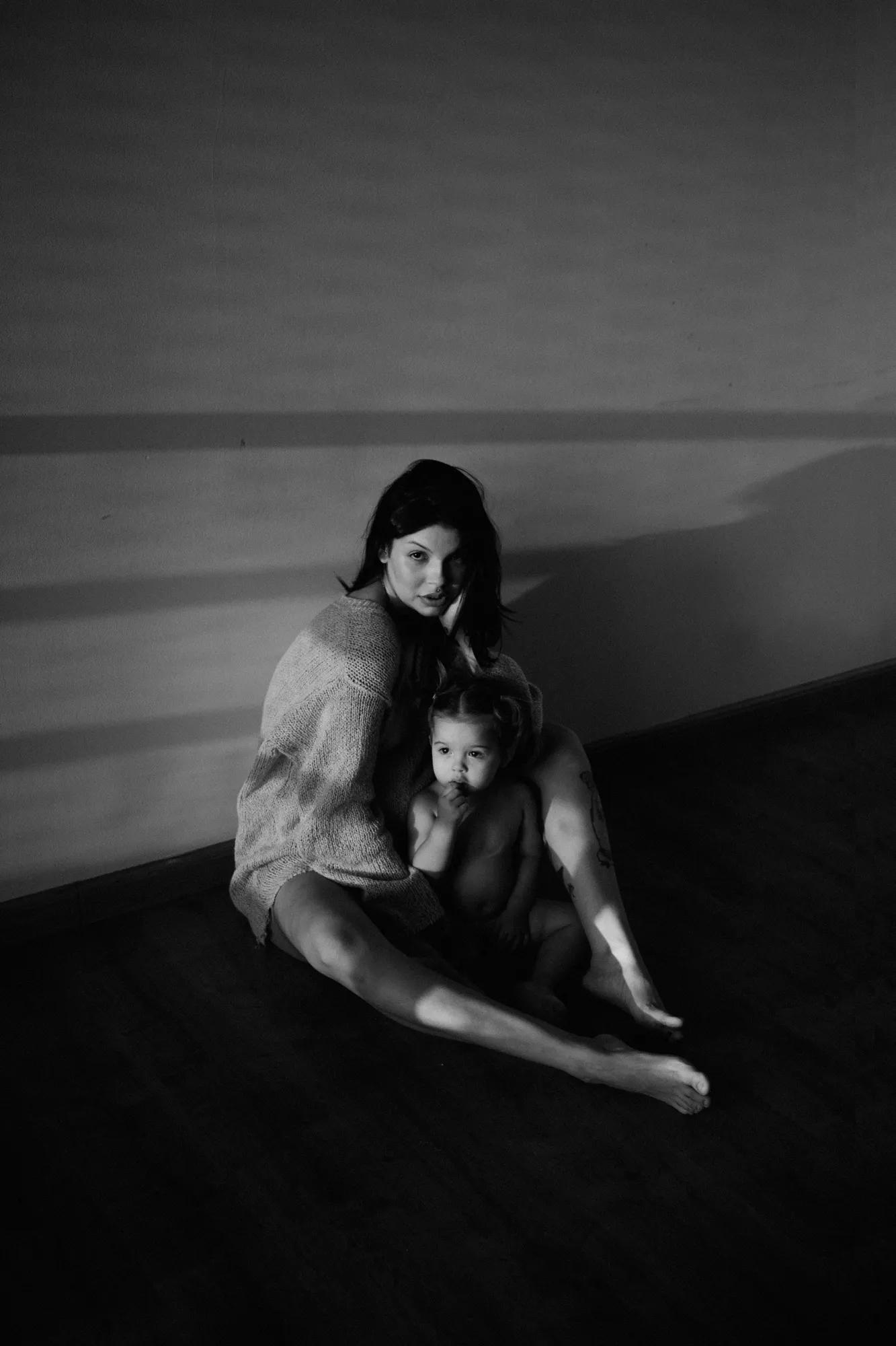 Pregnant woman sitting on the floor holding her oldest daughter. She is wearing a trashed sweater and looks at camera. shot on black and white