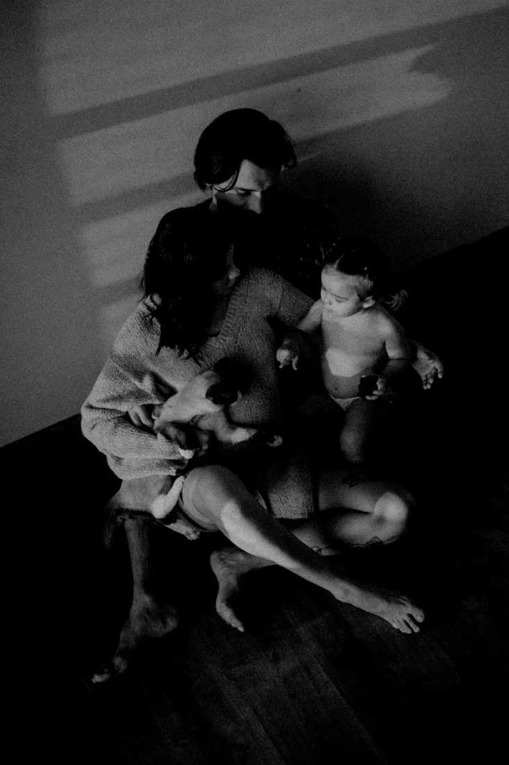 A family on sunlight. Father and pregnant mother holding their first child and cat. They are a sitting on the ground againt a wall. High angle shot. shot on black and white