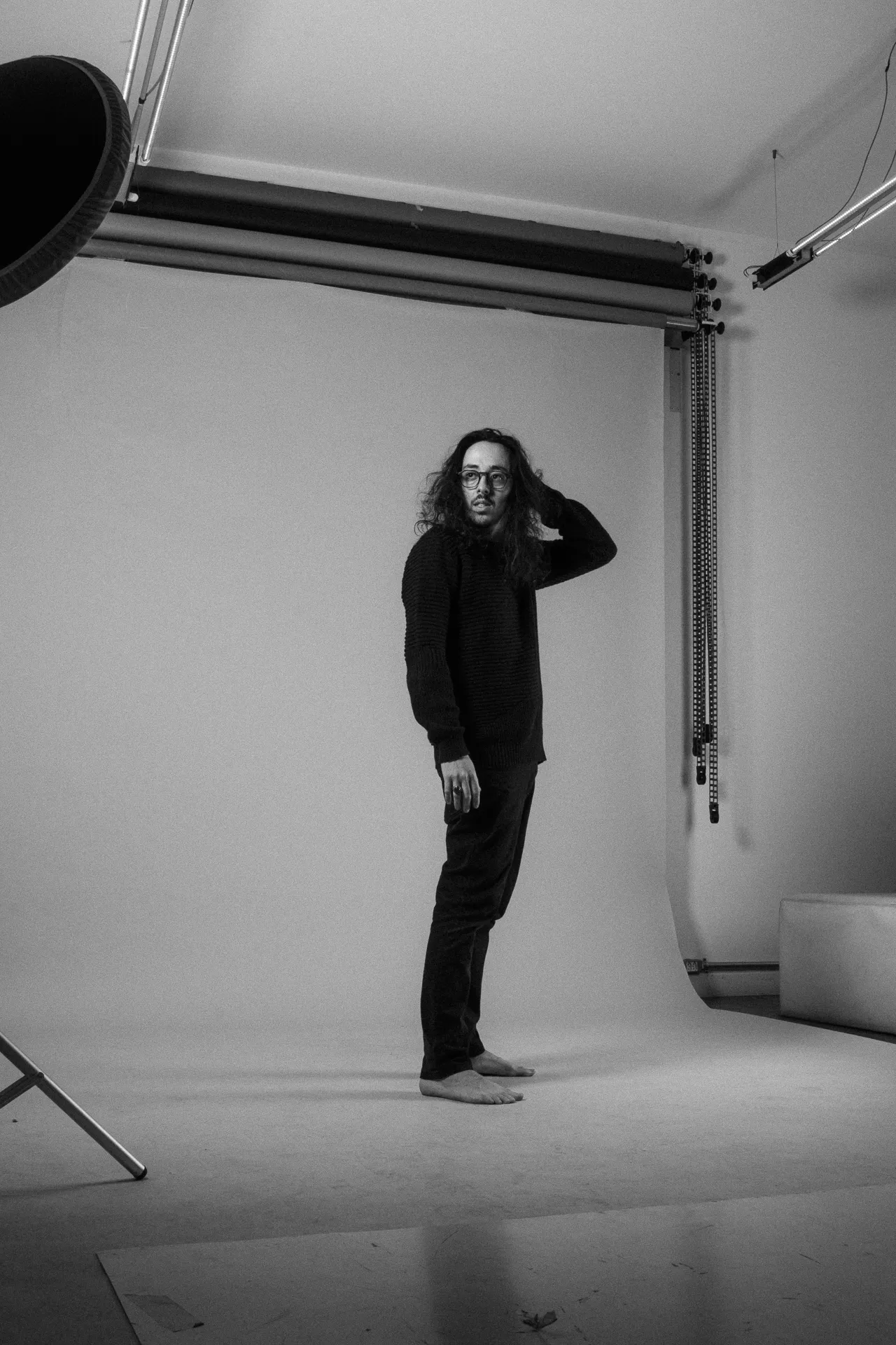 Foto do designer em preto e branco, em pé, em um estúdio, com cabelo solta e pés descalços