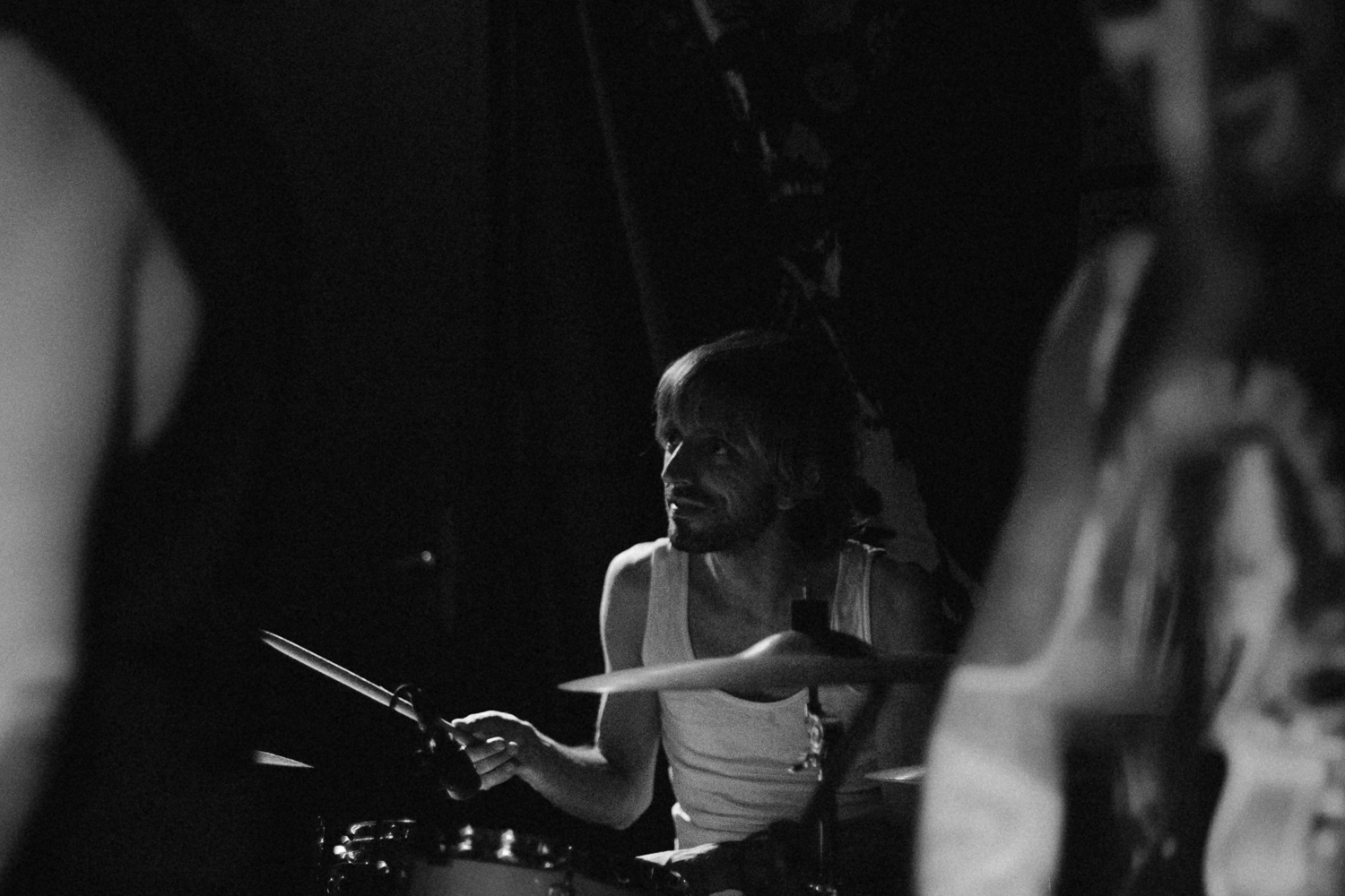 Black and White Live Concert Photography – Drummer in Performance by Rodrigo Baumgratz In this evocative black and white photograph by Rodrigo Baumgratz, a drummer is caught mid-performance, partially illuminated in a dim, textured atmosphere. With the other musicians blurred in the foreground, the focus lands squarely on the drummer's intent expression and rhythmic posture. The image captures the quiet intensity of live music from an insider's perspective — where noise meets stillness, and the beat becomes the breath. Ideal for portfolios centered on concert photography, music culture, or moody monochrome visual narratives. 