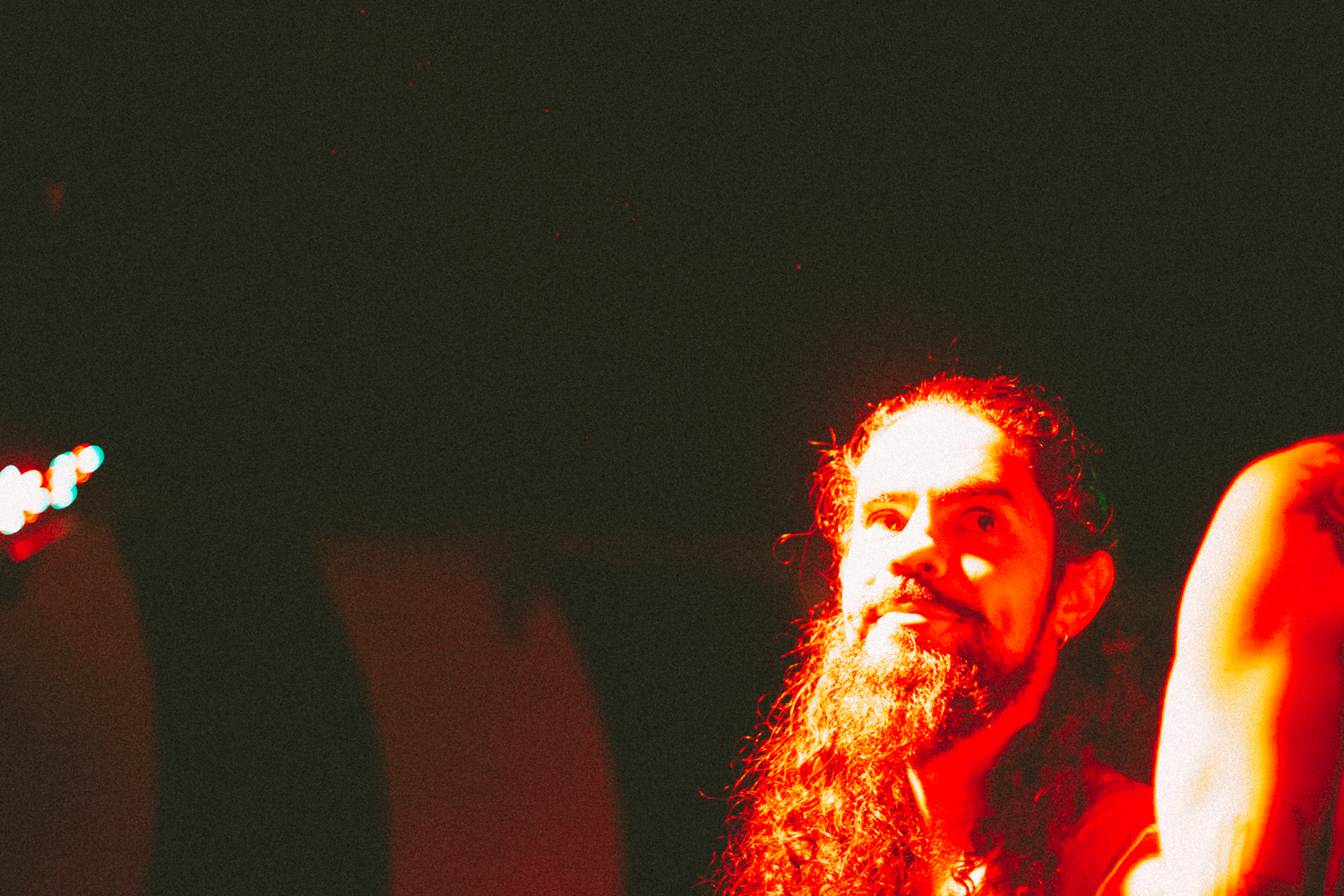 Expressive Concert Portrait – Red Stage Lighting on Performer by Rodrigo Baumgratz In this striking concert portrait by Rodrigo Baumgratz, a performer emerges from the darkness, bathed in bold red light. His long hair, full beard, and serene gaze evoke an intense, almost mythic presence. The use of extreme contrast and grainy texture adds a raw analog feel, perfect for portfolios exploring experimental music photography, emotional intensity, or alternative stage aesthetics. This image captures not only a performer, but the electrified silence between two songs.