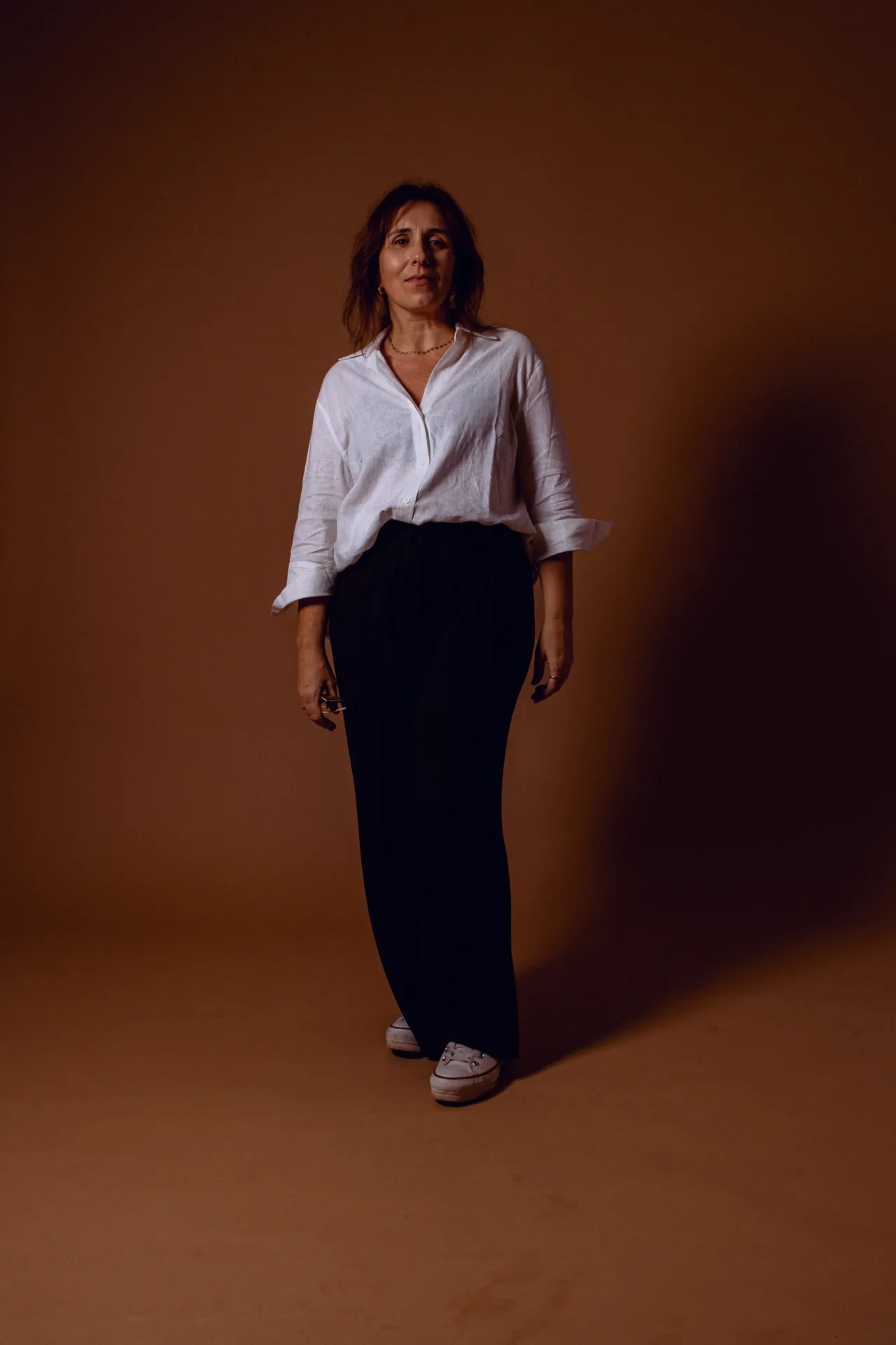 A woman with a white blouse and black pants on a brown background. She is redhead, mature and seams tired but in control