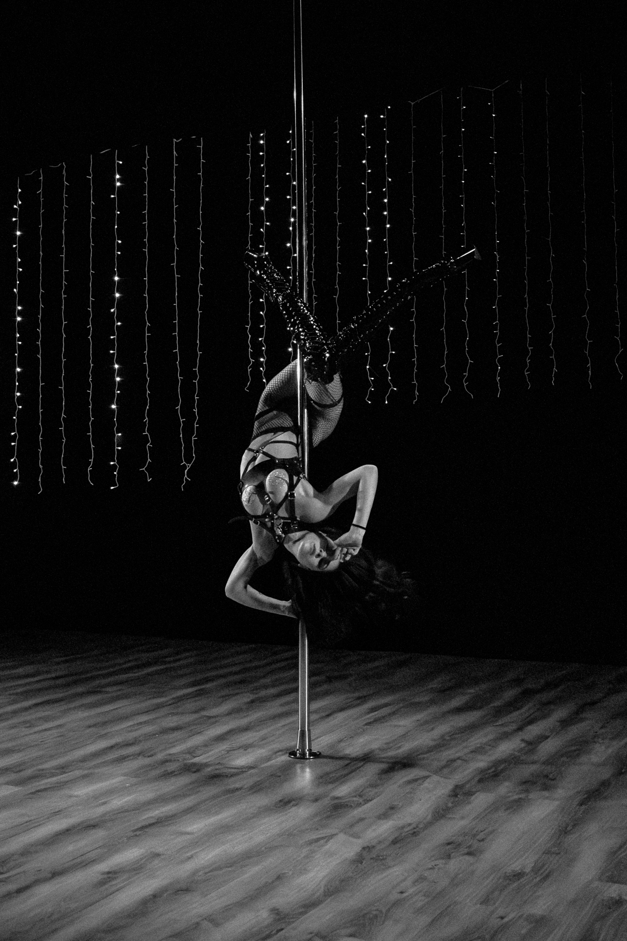 Black and White Pole Dance Portrait – Intimate Studio Photography by Rodrigo Baumgratz This black and white photograph by Rodrigo Baumgratz captures a powerful moment of pole dance performance. Suspended upside down with fluid strength, the performer is framed by a minimalist studio environment adorned with delicate vertical string lights. The high-contrast lighting emphasizes movement, texture, and body control, offering a striking visual of empowerment and grace. This image is ideal for portfolios focused on dance photography, performance art, studio portraiture, and gendered expression through movement. Keywords: pole dance photography, studio portrait, black and white dance image, Rodrigo Baumgratz, performance art photo, strength and grace.