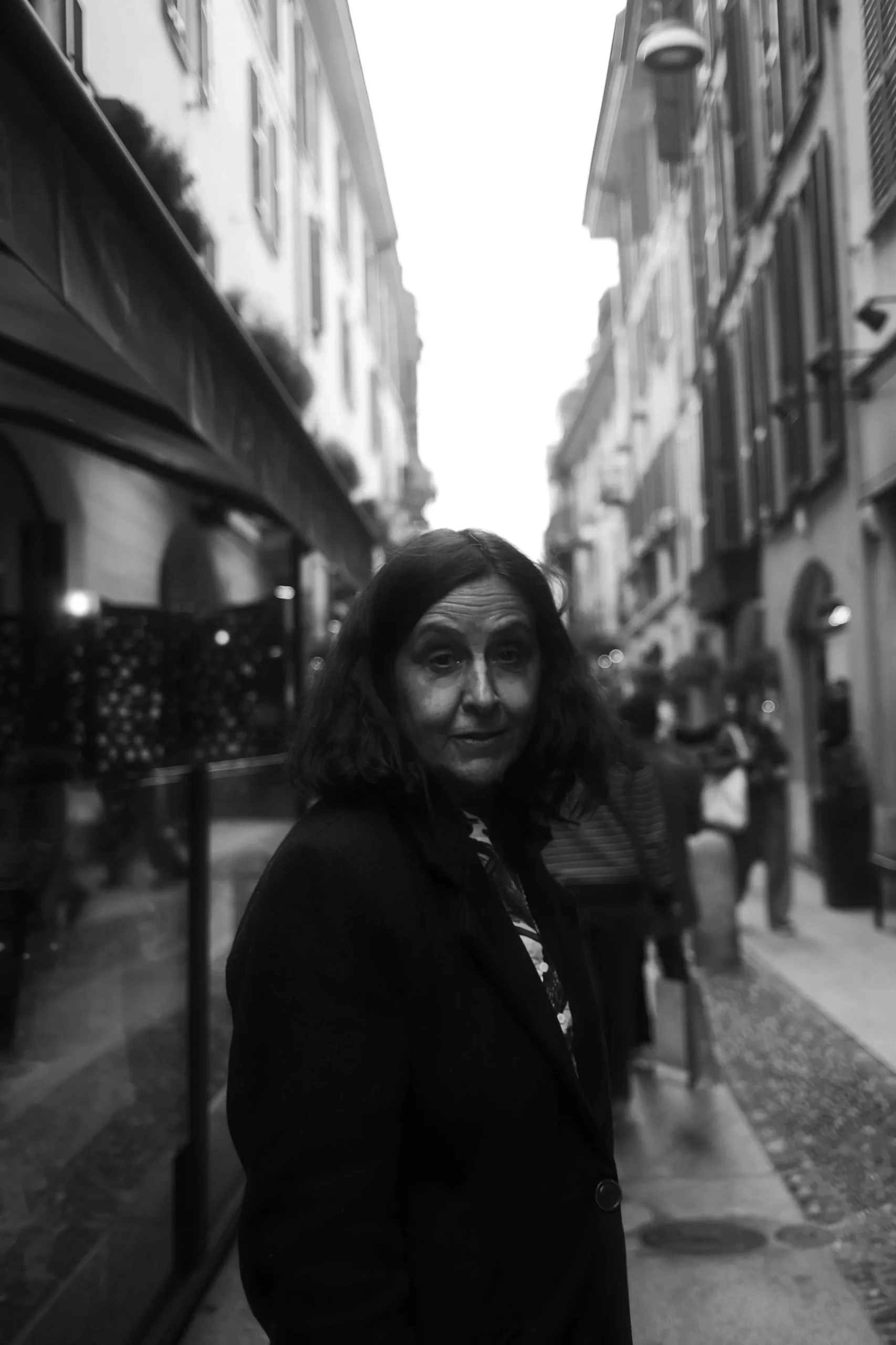Candid Portrait in Urban Passage – Black and White Photography by Rodrigo Baumgratz Captured on a narrow street in Milan, this black and white portrait centers on a solitary figure amidst an active urban backdrop. Rodrigo Baumgratz balances emotional subtlety with compositional restraint, highlighting human presence within the flow of city life. Ideal for editorial portraits, documentary-style campaigns, or projects exploring identity in public space.