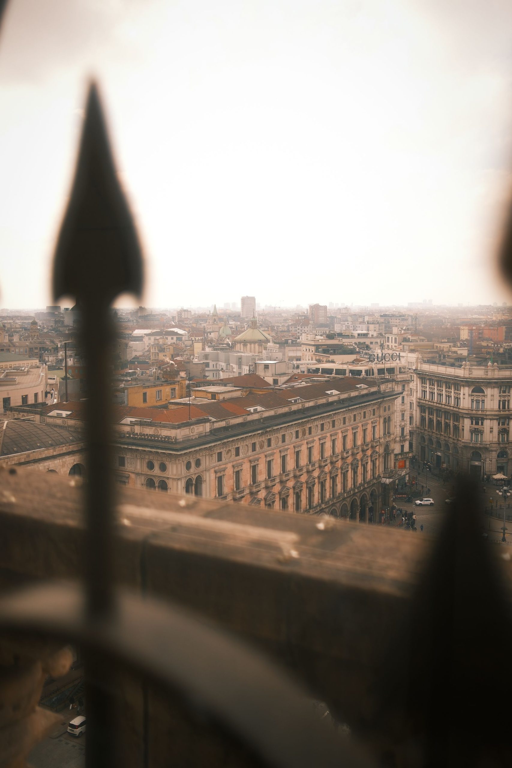 Framed Cityscape of Milan – View from Duomo Cathedral by Rodrigo Baumgratz Through the blurred iron spikes of the cathedral terrace, Milan unfolds in soft light and muted tones. Rodrigo Baumgratz frames the city as both distant and intimate, capturing rooftops and facades through an almost cinematic veil. The haze and framing suggest a narrative of longing, memory, and perspective — ideal for portfolios exploring conceptual urban photography, architectural storytelling, or poetic travel imagery.