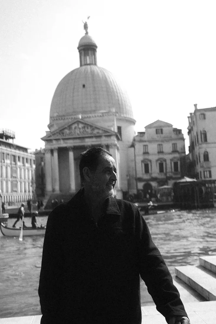 Editorial travel photography featured in a creative director portfolio, showing a man standing by a canal in Venice, Italy, with a historic domed church in the background. The image highlights natural daylight, cinematic composition, and environmental portraiture, emphasizing visual storytelling, art direction, and contemporary creative direction within a lifestyle and cultural context