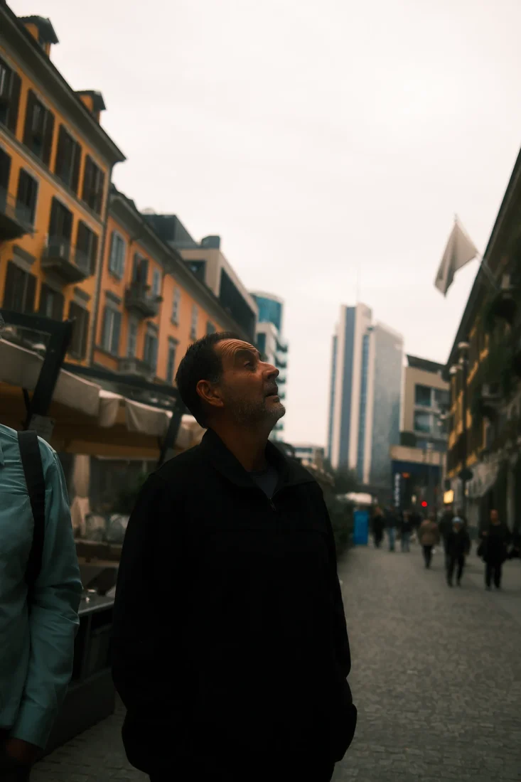 Documentary portrait of a man looking up on a Milan street, surrounded by buildings and people, exploring the contrast between everyday life and urban poetry.