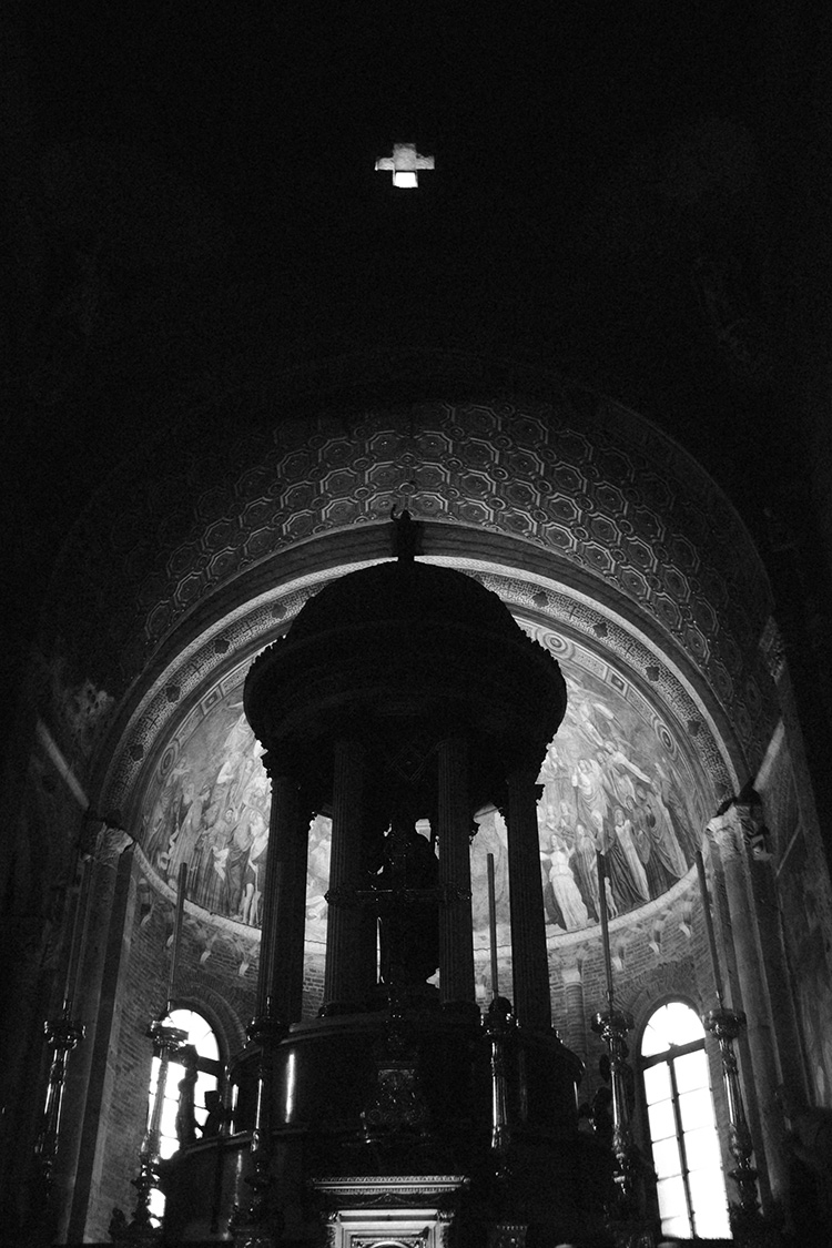 Sacred Light and Shadow – Black and White Church Interior Photography by Rodrigo Baumgratz. This striking monochrome image captures a dramatic play of light and darkness inside a historic church, with a cross-shaped skylight casting divine illumination over the richly detailed apse and altar. The high contrast reveals intricate frescoes and architectural elements while evoking a cinematic, almost mystical atmosphere. A fine example of visual storytelling through chiaroscuro, perfect for art direction, cultural editorials, and brands seeking depth and reverence in visual communication.