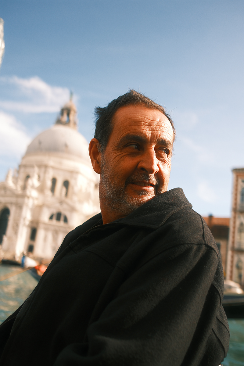 Editorial portrait photography featured in a creative director portfolio, capturing a middle-aged man in natural light on a boat in Venice, Italy. The composition combines cinematic framing, shallow depth of field, and warm color grading, showcasing visual storytelling, art direction, and lifestyle photography within a contemporary creative direction context