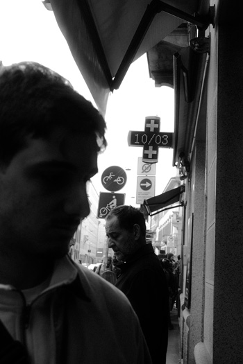 Street Scene in Milan – Layered Urban Portrait by Rodrigo Baumgratz Captured in stark black and white, this Milan street photograph by Rodrigo Baumgratz juxtaposes two figures caught in their own worlds. Signs, shadows, and urban noise crowd the frame, turning the composition into a visual allegory of disconnection. With strong contrast and raw textures, the photo embodies the essence of street photography — chaotic, visceral, and unapologetically real.