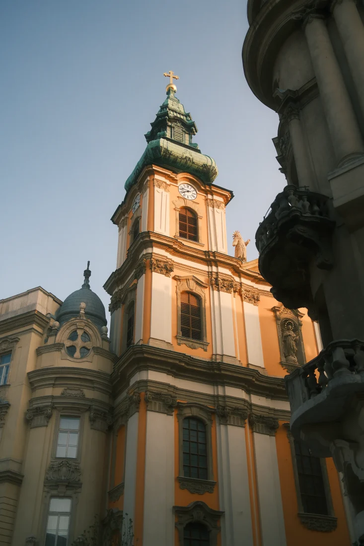 Architectural photograph of an ornate Baroque church in Budapest, captured in warm afternoon light with twin towers and green copper roofs. Creative director portfolio photography focused on historic architecture and visual composition.