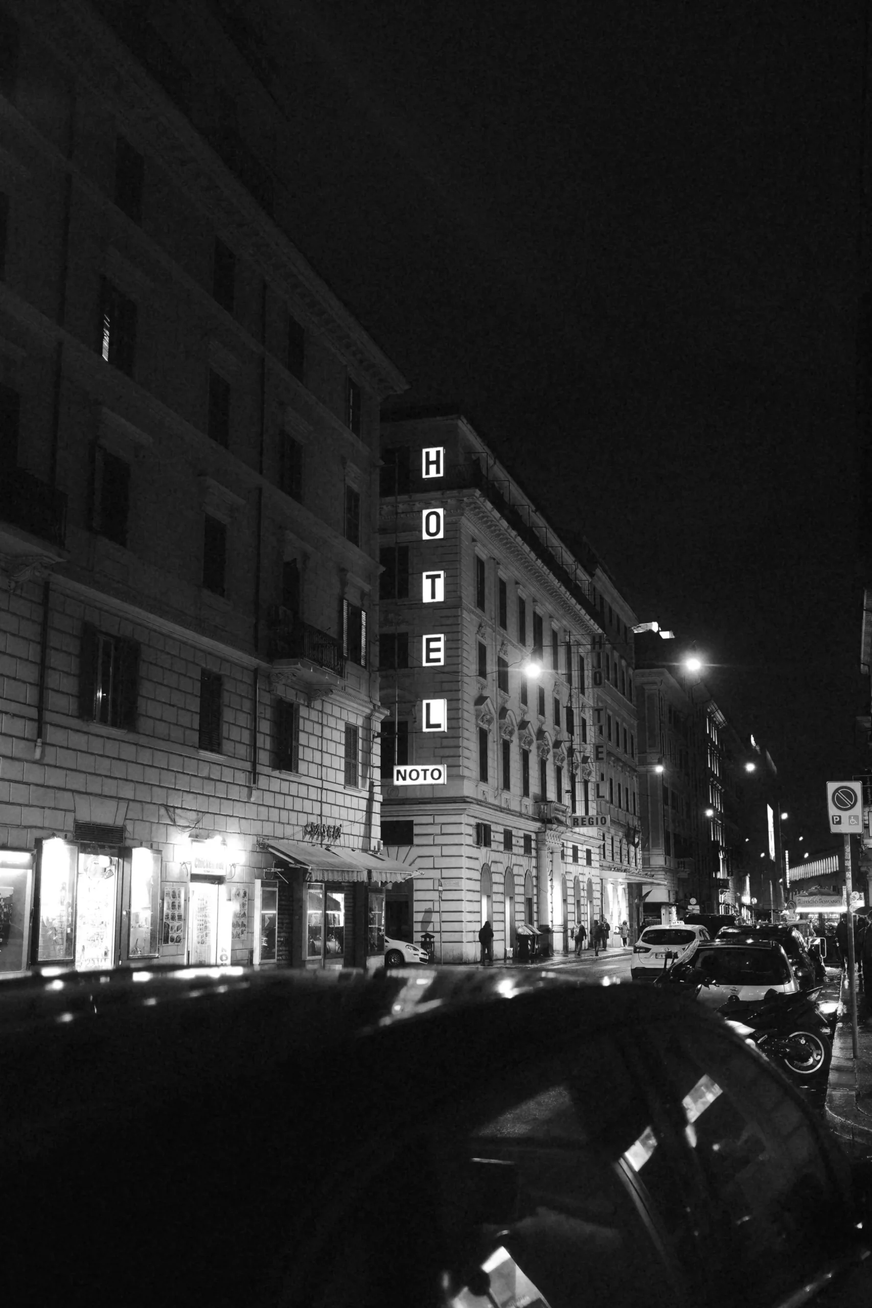 Black and white night photograph of Hotel Noto in Rome, Italy, framed by historic architecture, parked cars and illuminated street signs. An urban street photography image exploring nocturnal atmosphere, contrast, light and the cinematic character of Rome after dark.