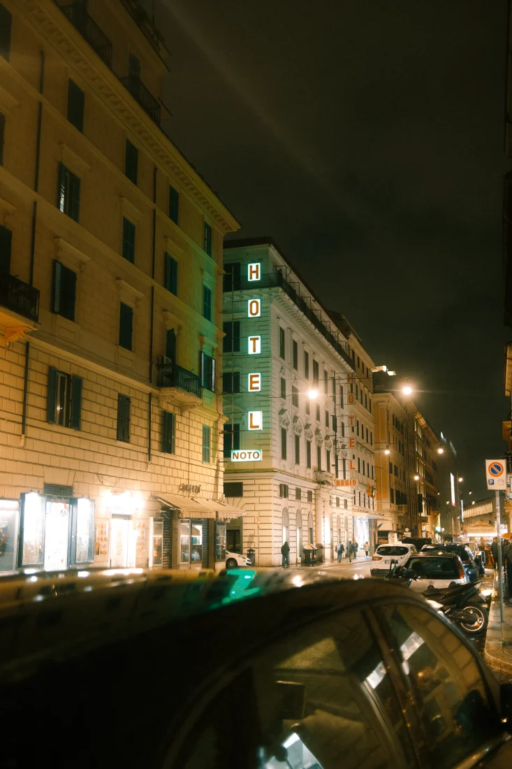 night photograph of Hotel Noto in Rome, Italy, framed by historic architecture, parked cars and illuminated street signs. An urban street photography image exploring nocturnal atmosphere, contrast, light and the cinematic character of Rome after dark.